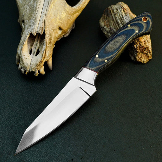 A fixed-blade knife with a satin-finished silver blade and a dark blue marbled handle, displayed diagonally on a dark textured surface next to a bone skull and a piece of wood.