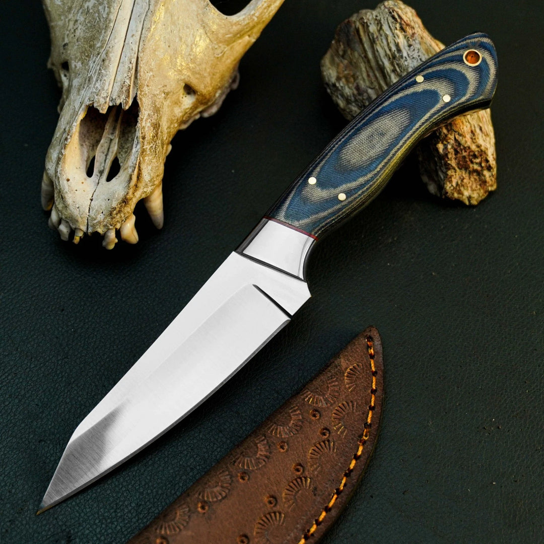 A fixed-blade knife with a satin-finished silver blade and a blue-gray wood handle with brass pins, displayed diagonally on a dark textured surface next to a brown leather sheath, a deer skull, and driftwood.