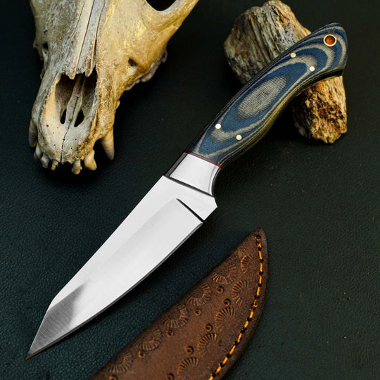 A fixed-blade knife with a satin-finished silver blade and a blue-gray wood handle with brass pins, displayed diagonally on a dark textured surface next to a brown leather sheath, a deer skull, and driftwood.