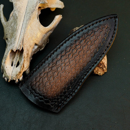 A dark brown textured leather knife sheath with black edges, displayed on a dark surface next to a weathered animal skull.