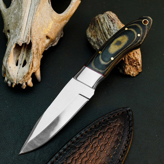 A fixed-blade knife with a satin-finished silver steel blade and a dark wood handle featuring a black, brown, and gold marbled pattern, displayed on a dark textured surface next to a leather sheath and a bone skull prop.