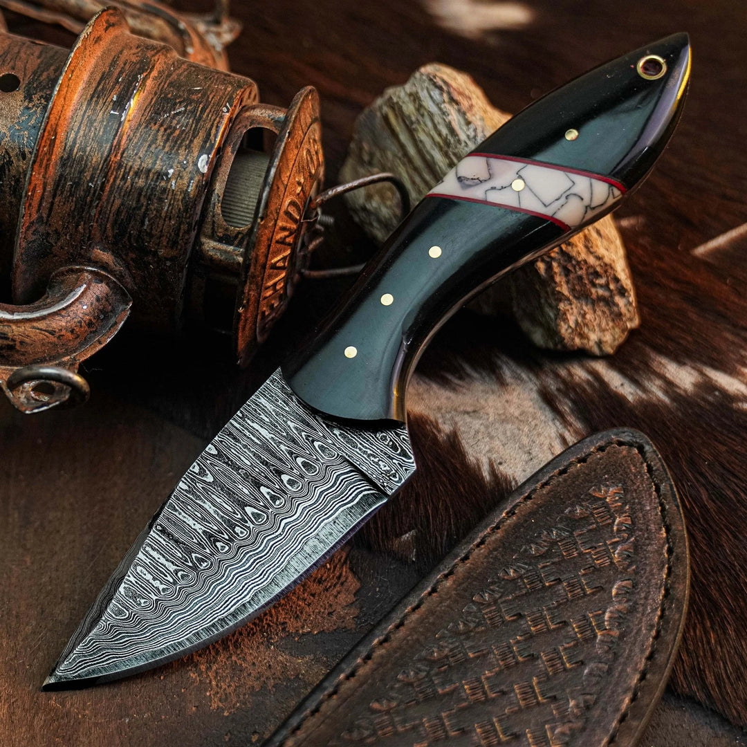 A fixed-blade knife with a patterned Damascus steel blade, a glossy black handle, and a white inlay with gold dots, displayed diagonally on a rustic background with a leather sheath, a wooden object, and rocks.