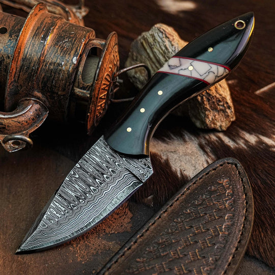 A fixed-blade knife with a patterned Damascus steel blade, a glossy black handle, and a white inlay with gold dots, displayed diagonally on a rustic background with a leather sheath, a wooden object, and rocks.