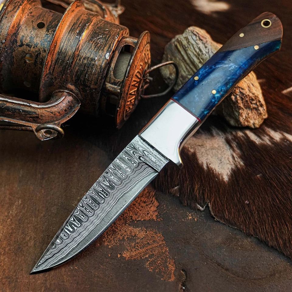 A custom fixed-blade knife with a Damascus steel blade and a blue marbled handle, displayed diagonally on a rustic background with a vintage fishing reel and cowhide.