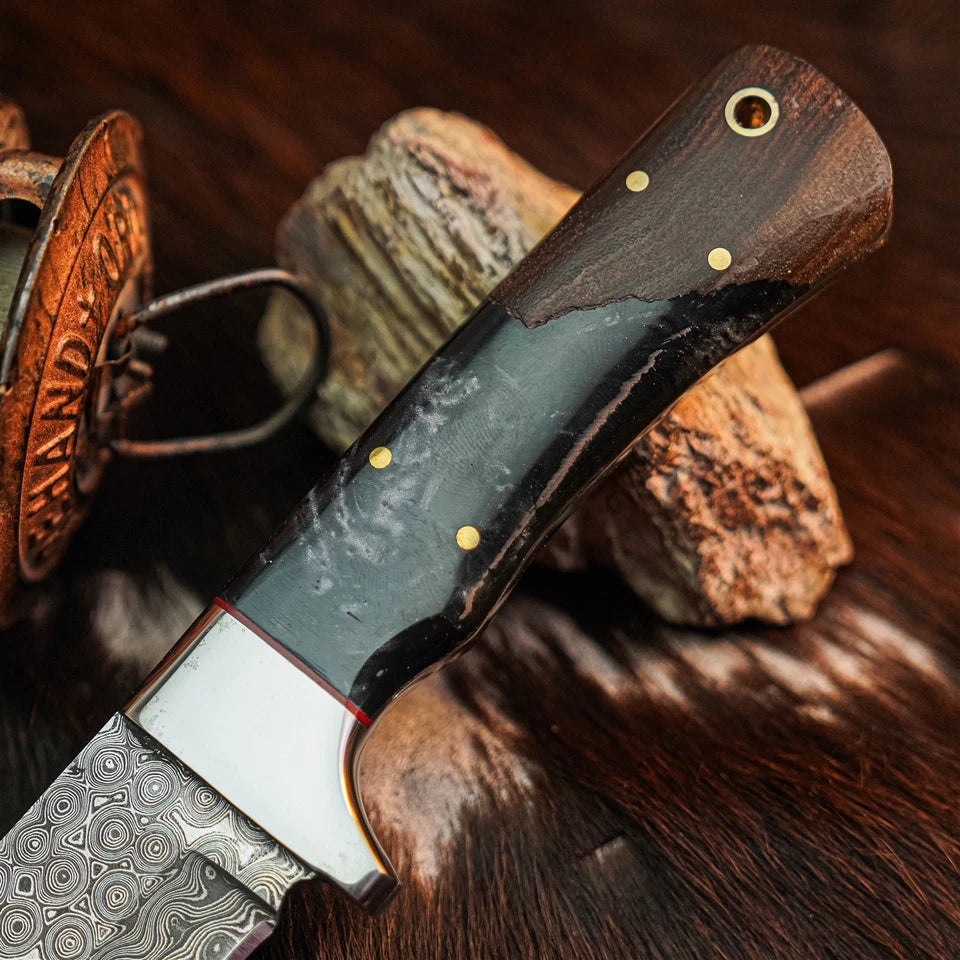 A close-up of a handcrafted knife with a Damascus steel blade, a dark wood handle, and a black inlay, resting on a textured brown fur surface with rustic wood props.