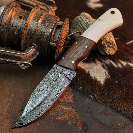 A fixed-blade knife with a patterned Damascus steel blade, a dark wood handle, and an ivory-colored bone handle section, displayed diagonally on a rustic brown surface next to a vintage lantern.
