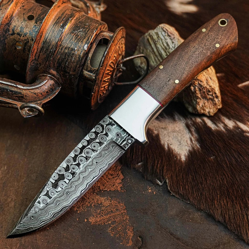 A fixed-blade knife with a patterned Damascus steel blade and a dark brown wooden handle, styled on a rustic background with a vintage fly rod and cowhide.
