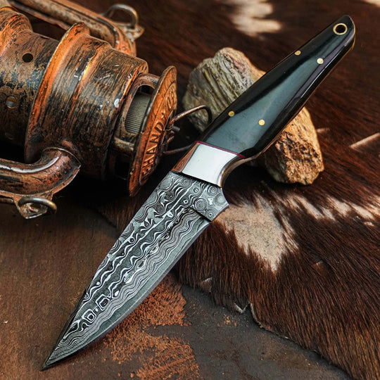 A fixed-blade knife with a patterned Damascus steel blade and a dark handle, displayed diagonally on a rustic brown hide with a vintage fishing reel prop.