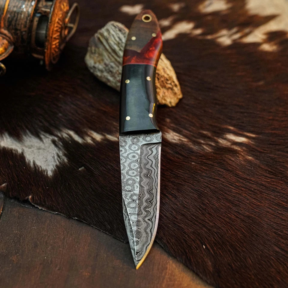A custom handmade knife with a Damascus steel blade and a carved wooden handle, displayed on a brown and white cowhide rug with rustic western props.