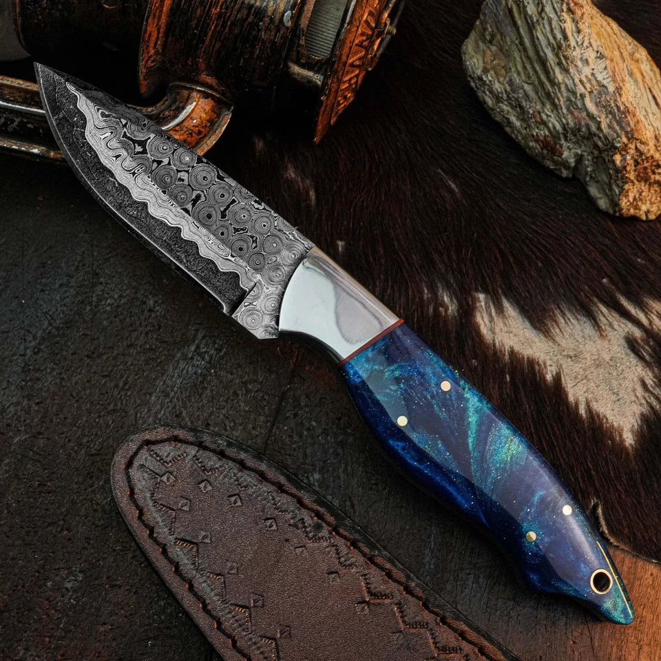 A fixed-blade knife with a patterned steel blade and a blue marbled resin handle, displayed diagonally on a dark textured surface with a brown leather sheath, fur, and wood props.