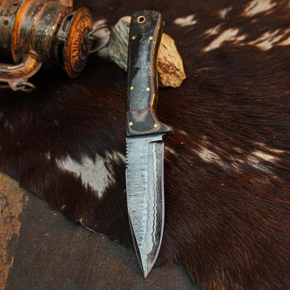 A handmade fixed-blade knife with a Damascus steel blade and a dark wood handle with brass pins, resting on a brown and white cowhide pelt with a rustic fishing reel prop in the background.