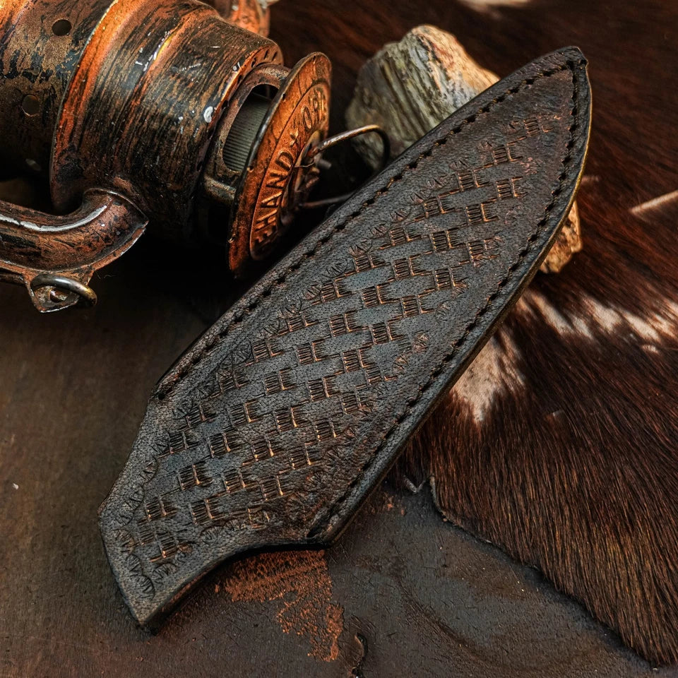 A dark brown leather knife sheath with an embossed basketweave pattern and visible stitching, displayed diagonally on a rustic background with a vintage fishing reel and cowhide.