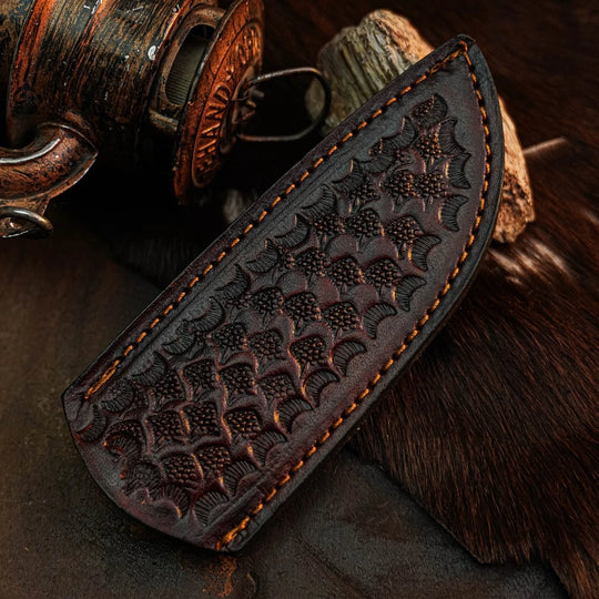 A dark brown leather sheath with an embossed fish-scale pattern and orange-brown contrast stitching, displayed on a dark textured surface with rustic props.