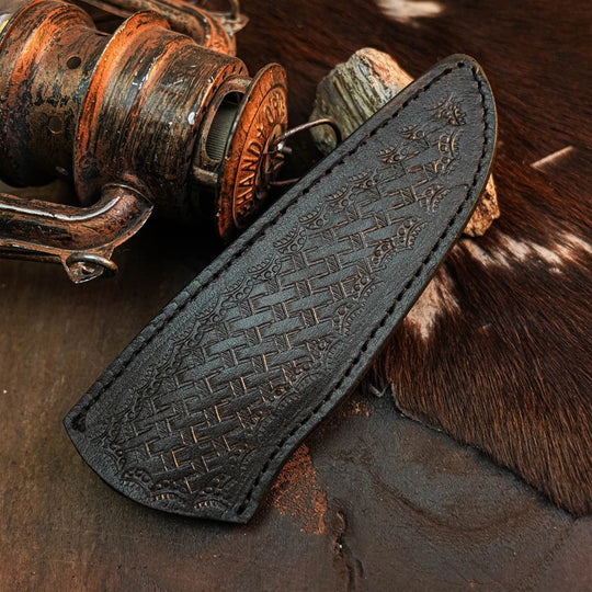 A black leather knife sheath with an embossed basketweave pattern and visible stitching, displayed diagonally on a rustic dark surface next to a vintage copper fishing reel and cowhide.