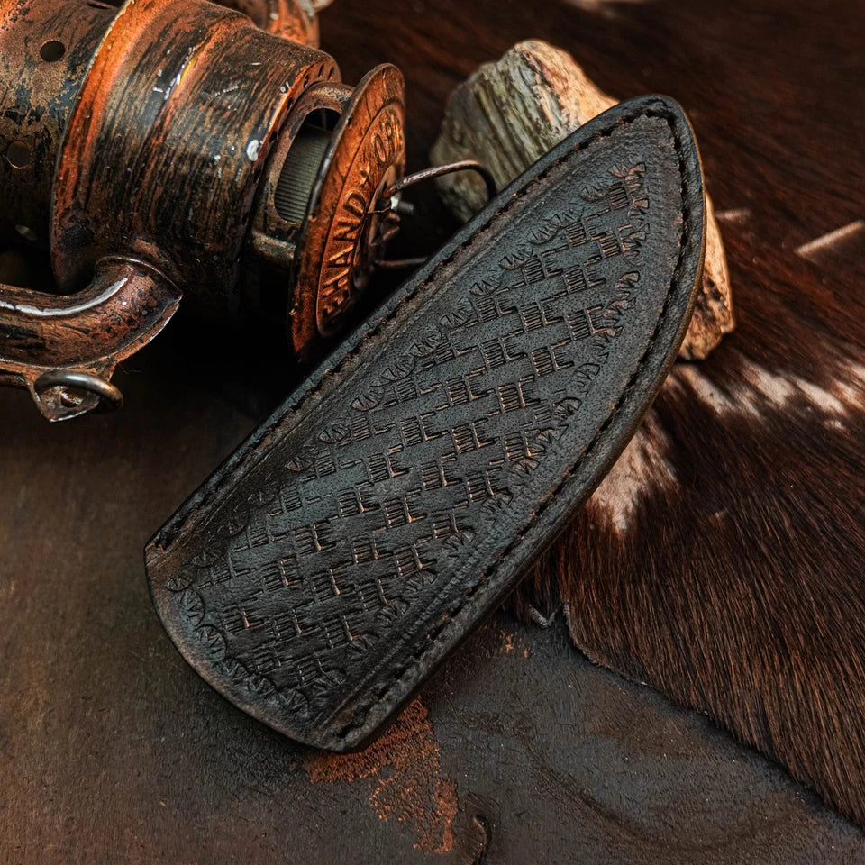 A dark brown leather sheath with an embossed basketweave pattern, shown resting on a rustic background with a vintage copper fishing reel and textured fur.