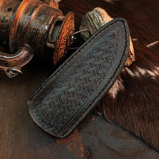 A dark brown leather sheath with an embossed basketweave pattern, shown resting on a rustic background with a vintage copper fishing reel and textured fur.