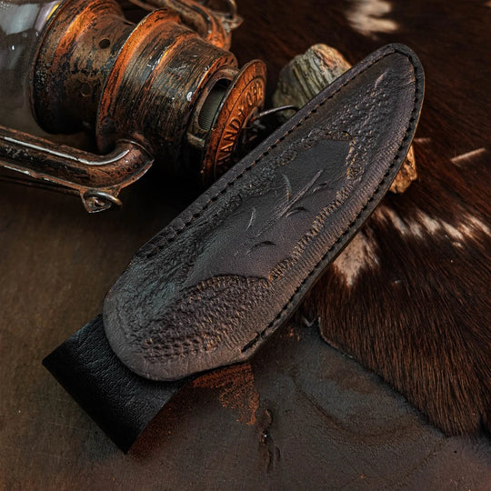 A dark brown leather knife sheath with an embossed floral design, displayed diagonally on a rustic background with an antique lantern and cowhide.
