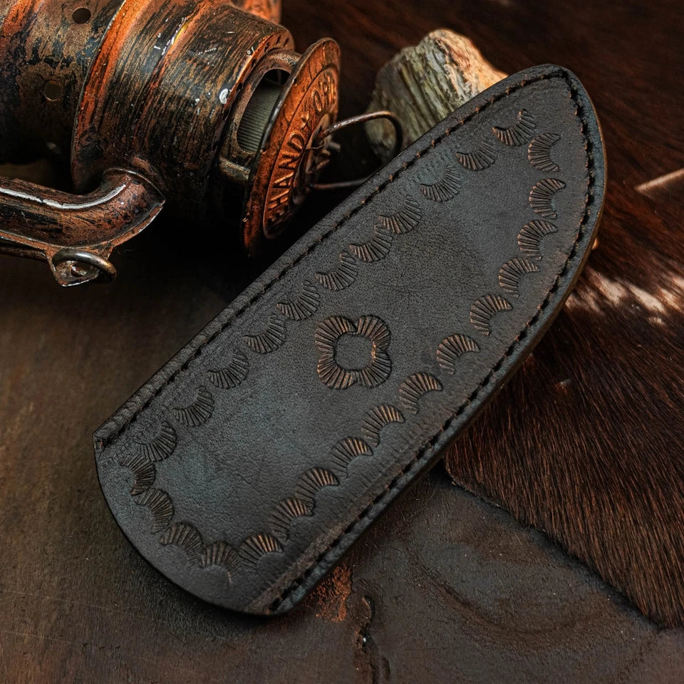 A dark brown leather knife sheath with hand-stitched edges and embossed floral patterns, displayed on a rustic dark wooden surface next to an antique fishing reel.