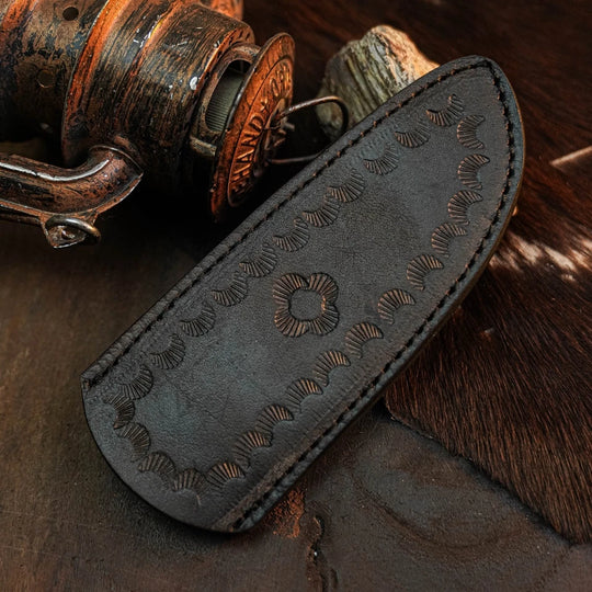 A dark brown leather knife sheath with hand-stitched edges and embossed floral patterns, displayed on a rustic dark wooden surface next to an antique fishing reel.