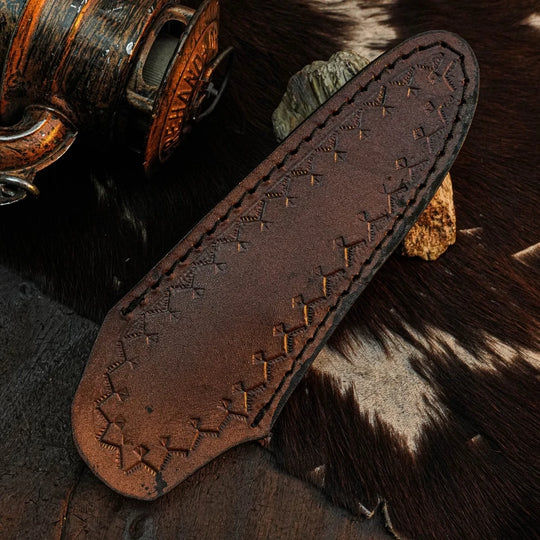 A brown leather knife sheath with decorative stamped geometric patterns and stitched edges, displayed on a rustic dark wood and cowhide surface.