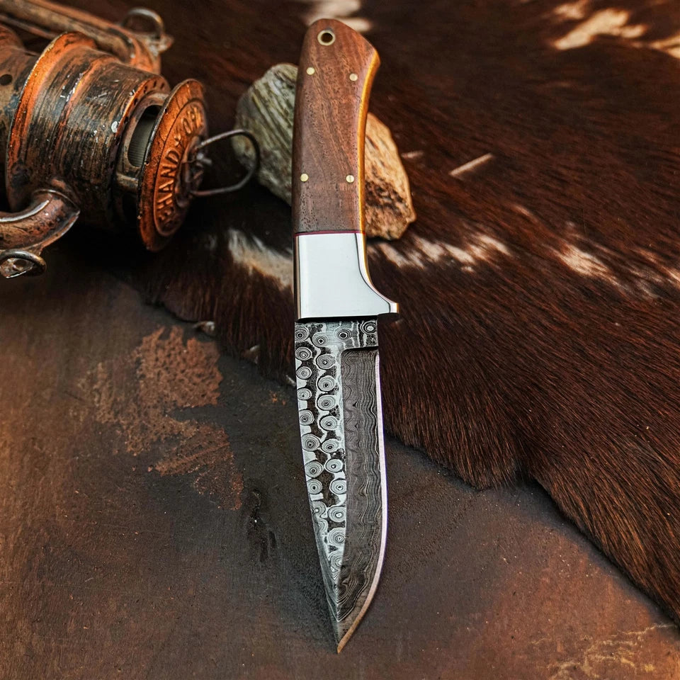 A fixed-blade knife with a patterned steel blade, a silver bolster, and a brown wooden handle, displayed on a rustic brown surface with a cowhide and an antique lantern in the background.
