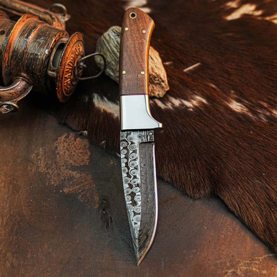 A fixed-blade knife with a patterned steel blade, a silver bolster, and a brown wooden handle, displayed on a rustic brown surface with a cowhide and an antique lantern in the background.