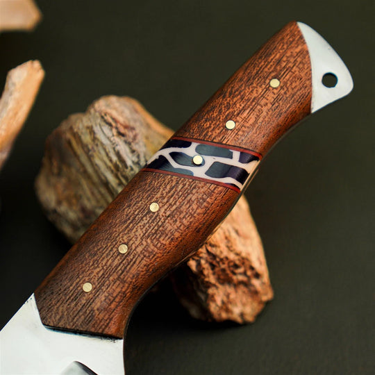A close-up of a knife handle made of rich brown wood with brass pins and a black, white, and red inlay, resting on rustic wood pieces against a dark background.
