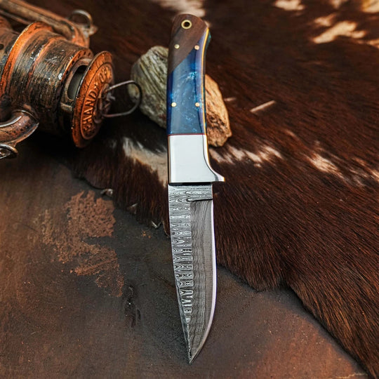 A custom handmade fixed-blade knife with a patterned Damascus steel blade and a blue marbled resin handle, displayed on a brown cowhide rug next to a rustic wooden object.