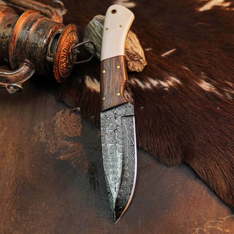 A fixed-blade knife with a patterned Damascus steel blade, a dark wood handle, and a cream-colored bone or horn bolster, displayed on a brown leather and cowhide surface next to a rustic wooden object.