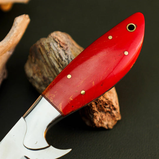 A close-up of a knife with a glossy red handle and brass pins, resting on rustic wood pieces against a dark background.