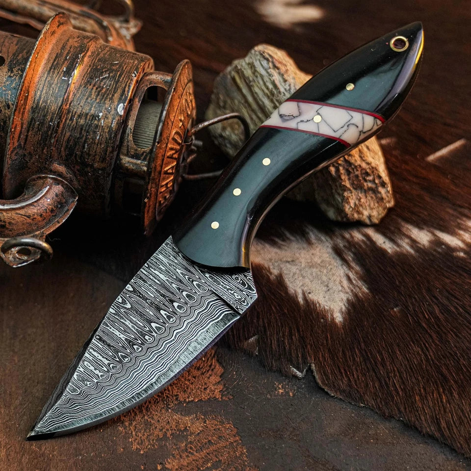 A fixed-blade knife with a patterned Damascus steel blade and a dark handle featuring a white inlay with gold dots, displayed on a rustic brown surface with a vintage reel and animal-hide texture.