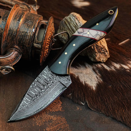 A fixed-blade knife with a patterned Damascus steel blade and a dark handle featuring a white inlay with gold dots, displayed on a rustic brown surface with a vintage reel and animal-hide texture.