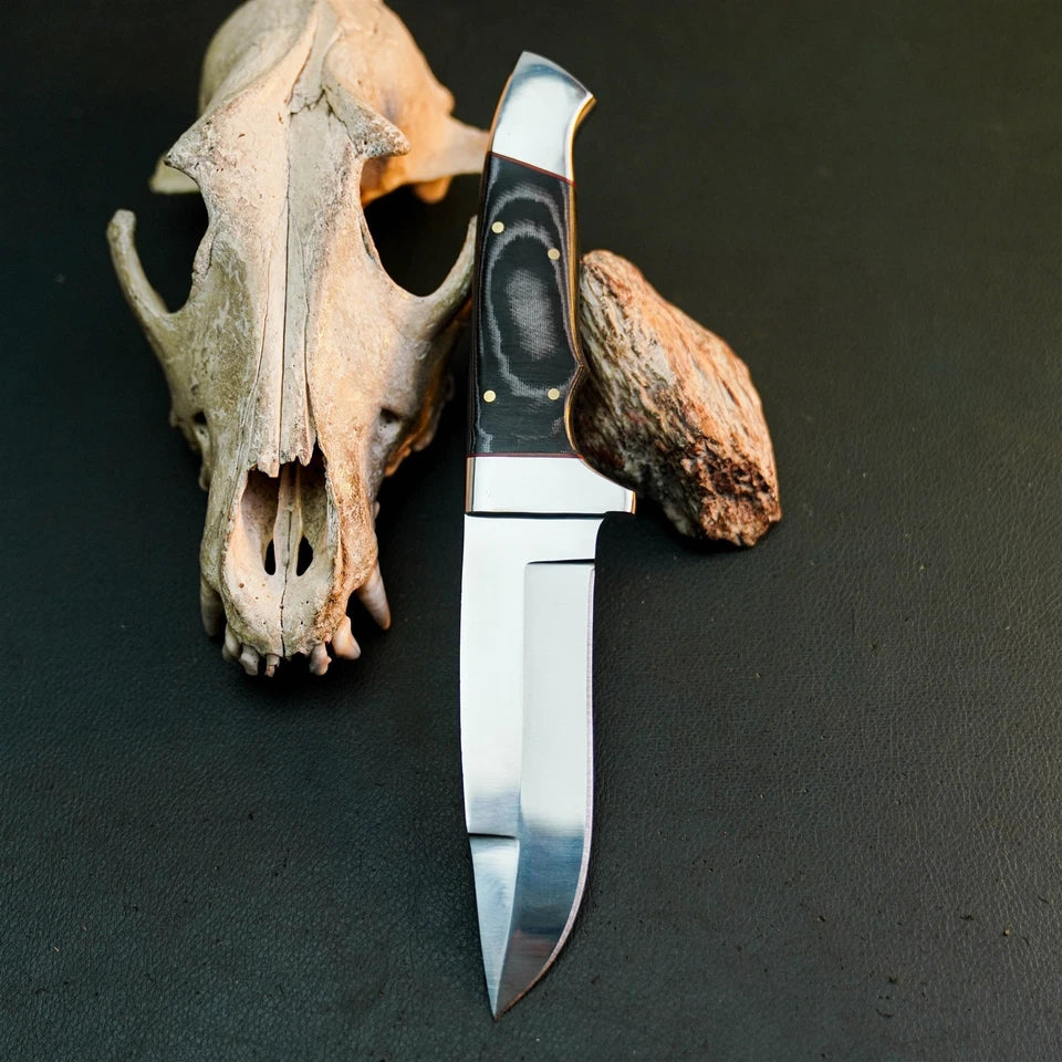 A fixed-blade knife with a polished silver blade and a black handle with gold accents, displayed vertically next to a deer skull and a small rock on a dark textured surface.