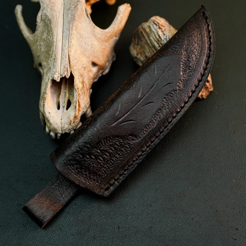 A dark brown leather knife sheath with embossed leaf detailing, shown with a matching leather knife handle loop, styled on a dark textured surface with a deer skull and wood props in the background.