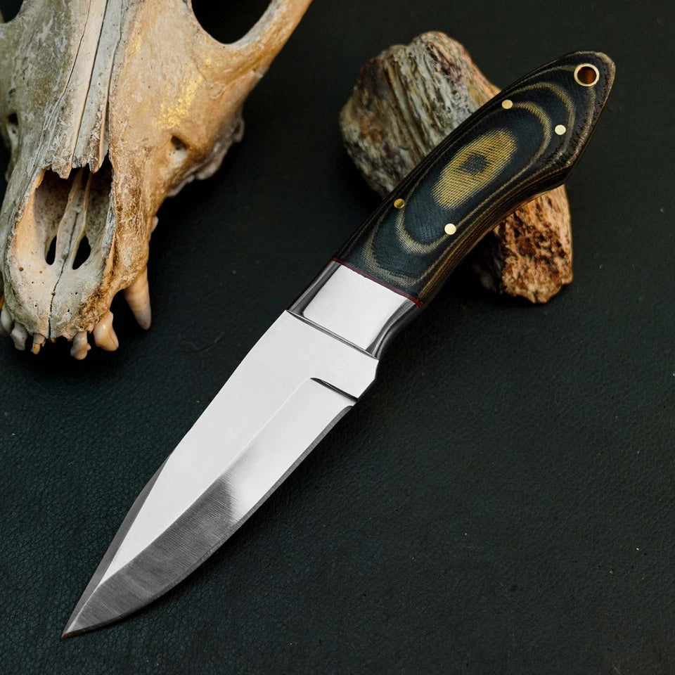 A fixed-blade knife with a satin-finished silver blade and a dark handle featuring a circular black, brown, and gold pattern, displayed diagonally on a dark textured surface next to a skull and rock.