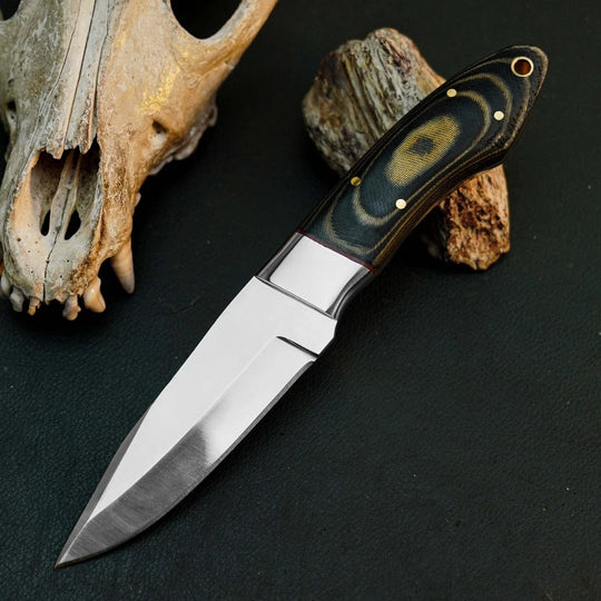 A fixed-blade knife with a satin-finished silver blade and a dark handle featuring a circular black, brown, and gold pattern, displayed diagonally on a dark textured surface next to a skull and rock.