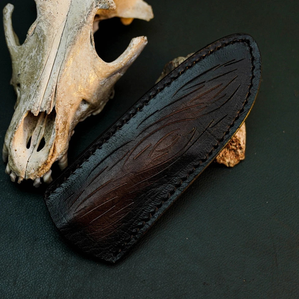 A dark brown leather sheath with engraved leaf-like patterns and visible stitching, displayed on a dark background next to an animal skull and small rock.