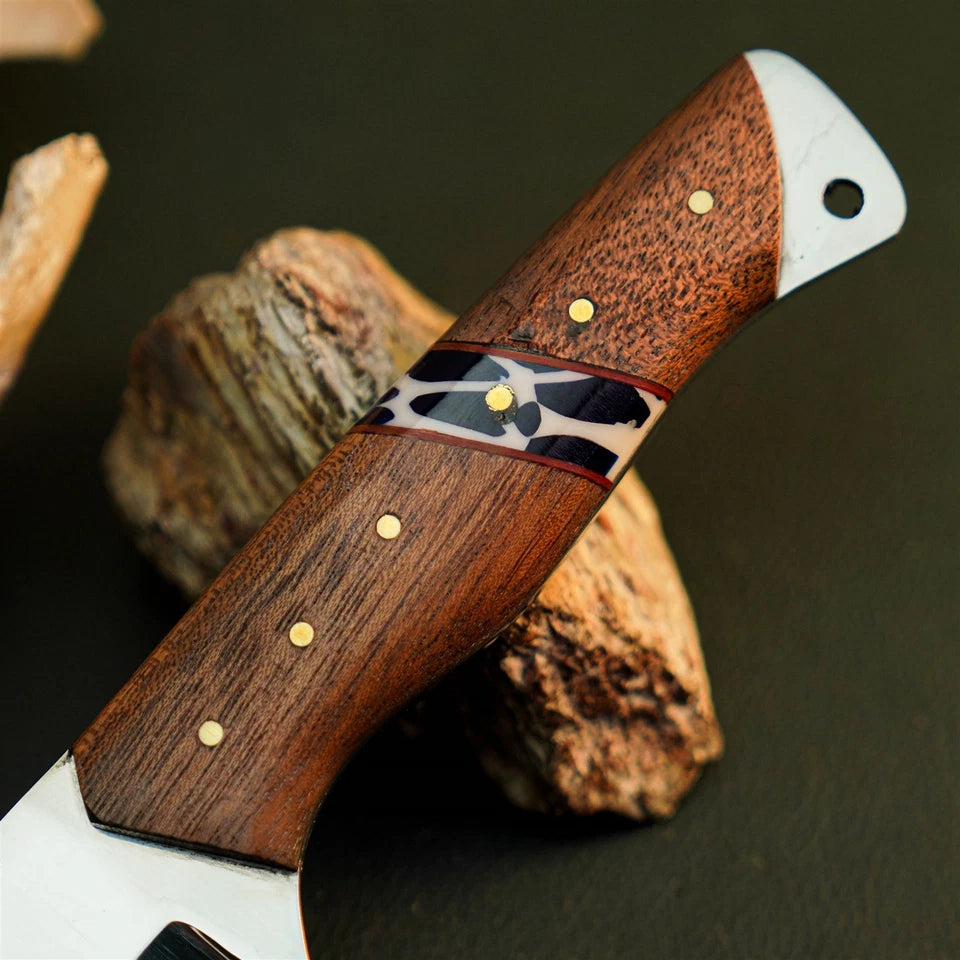 A close-up of a handcrafted knife with a rich brown wooden handle, gold brass pins, and a black-and-white inlay, resting on rustic wood pieces against a dark background.