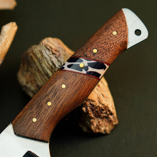 A close-up of a handcrafted knife with a rich brown wooden handle, gold brass pins, and a black-and-white inlay, resting on rustic wood pieces against a dark background.