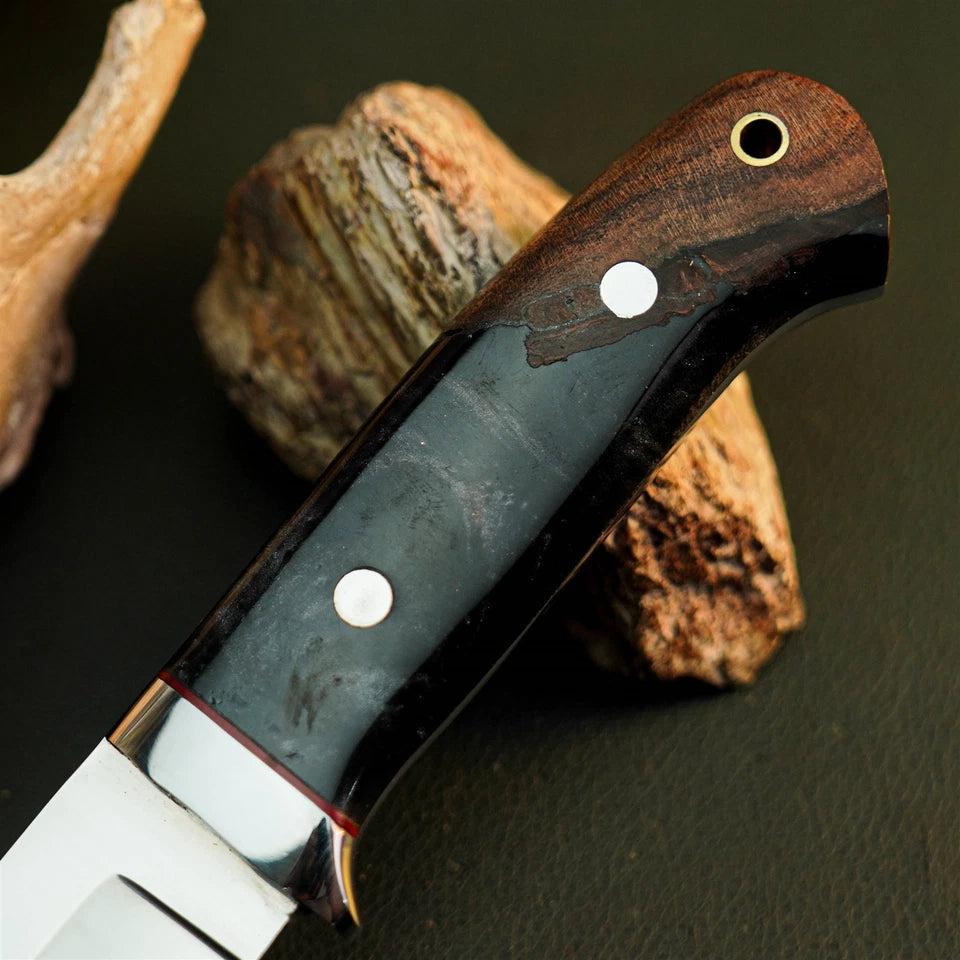 A close-up of a knife handle with a dark wood and black inlay, secured with silver pins, resting on natural driftwood against a dark background.