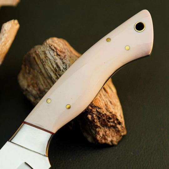 A close-up of a knife handle made of pale pink bone or horn, secured with brass pins, resting on a piece of wood against a dark background.