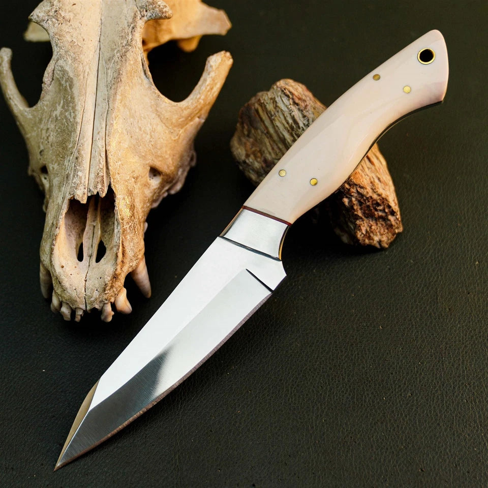 A fixed-blade knife with a polished silver blade and a light beige handle with brass pins, displayed diagonally on a dark textured surface next to a deer skull and a piece of wood.