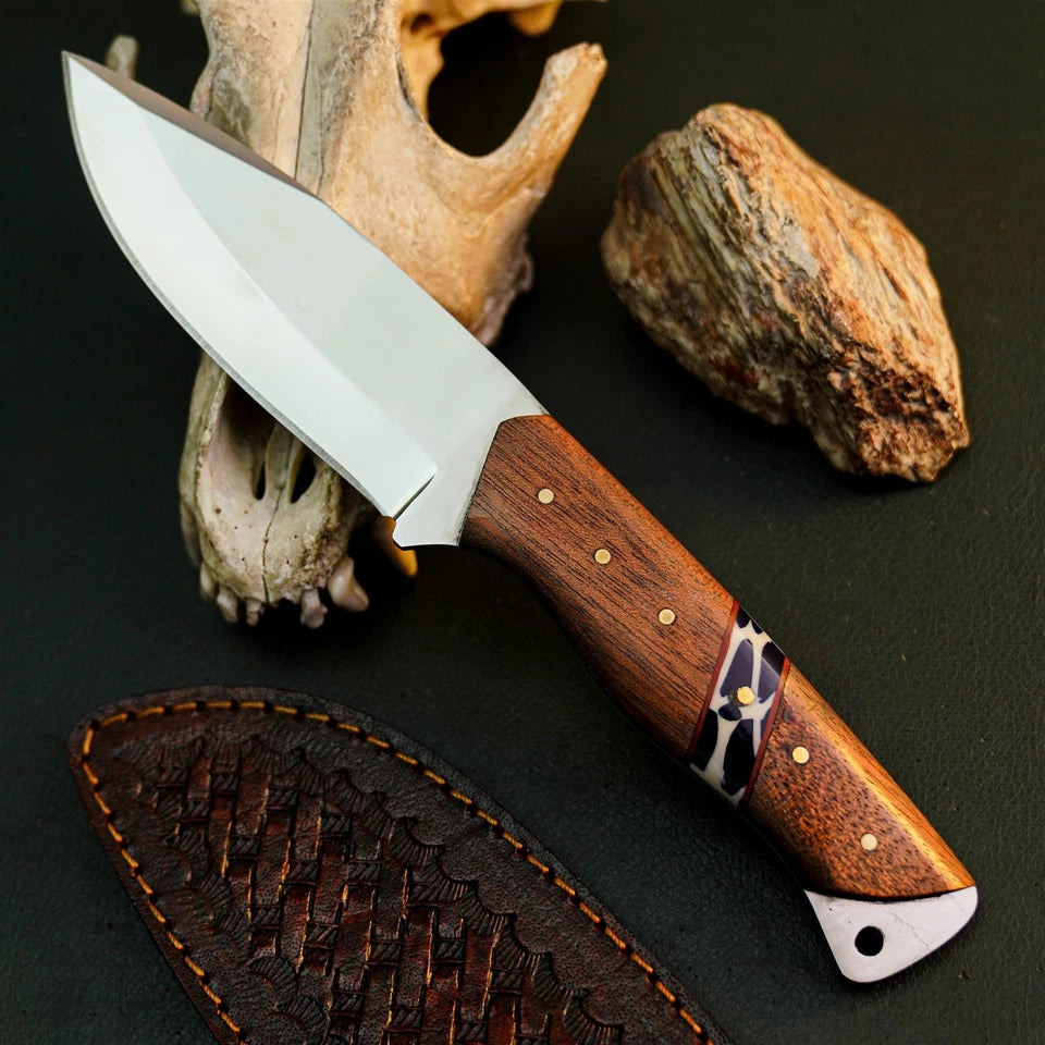 A fixed-blade knife with a polished silver blade and a brown wood handle with brass pins and a patterned inlay, displayed diagonally on a dark textured surface with a leather sheath, a bone fragment, and a rock.