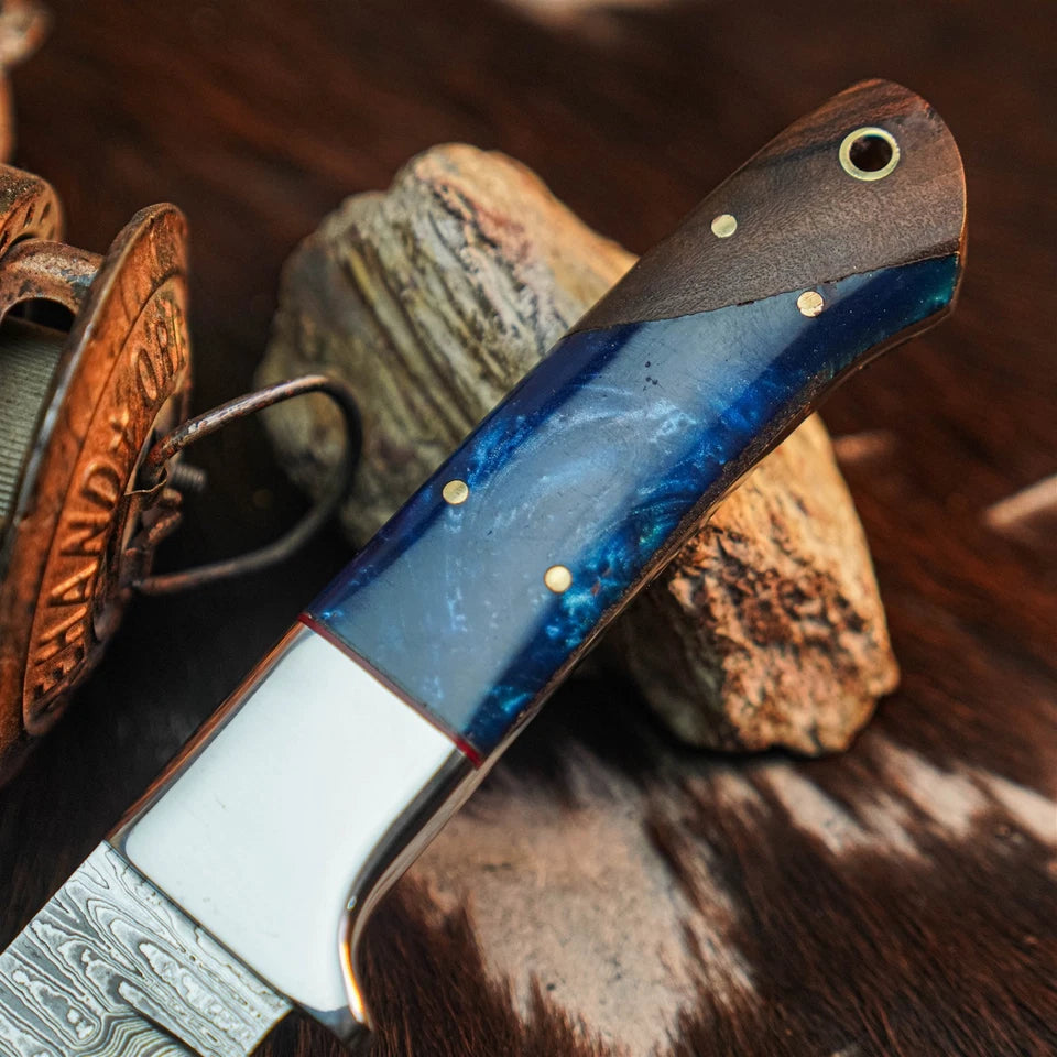 A close-up of a custom knife with a blue and white marbled handle, brass pins, and a Damascus steel blade, resting on rustic wood and cowhide.