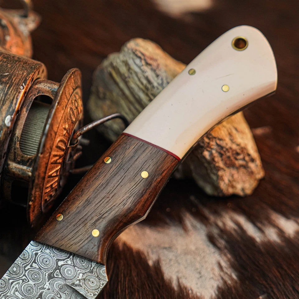 A close-up of a handcrafted knife with a patterned steel blade, a dark wood handle, and an ivory-colored end piece, resting on rustic wood and fur.