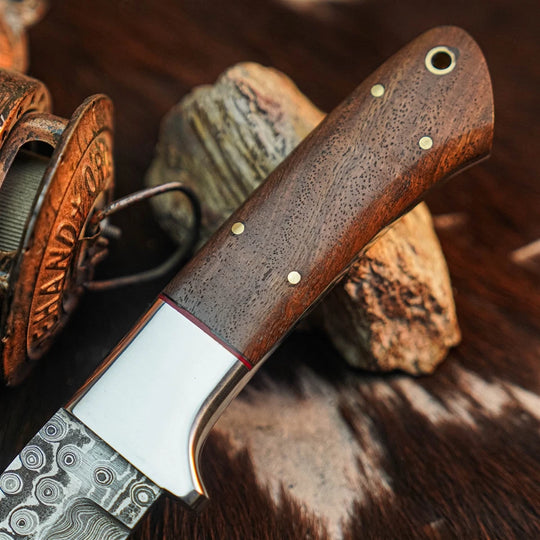 A close-up of a handcrafted knife with a dark brown wood handle, brass pins, and a patterned steel blade, resting on rocks and fur against a rustic background.