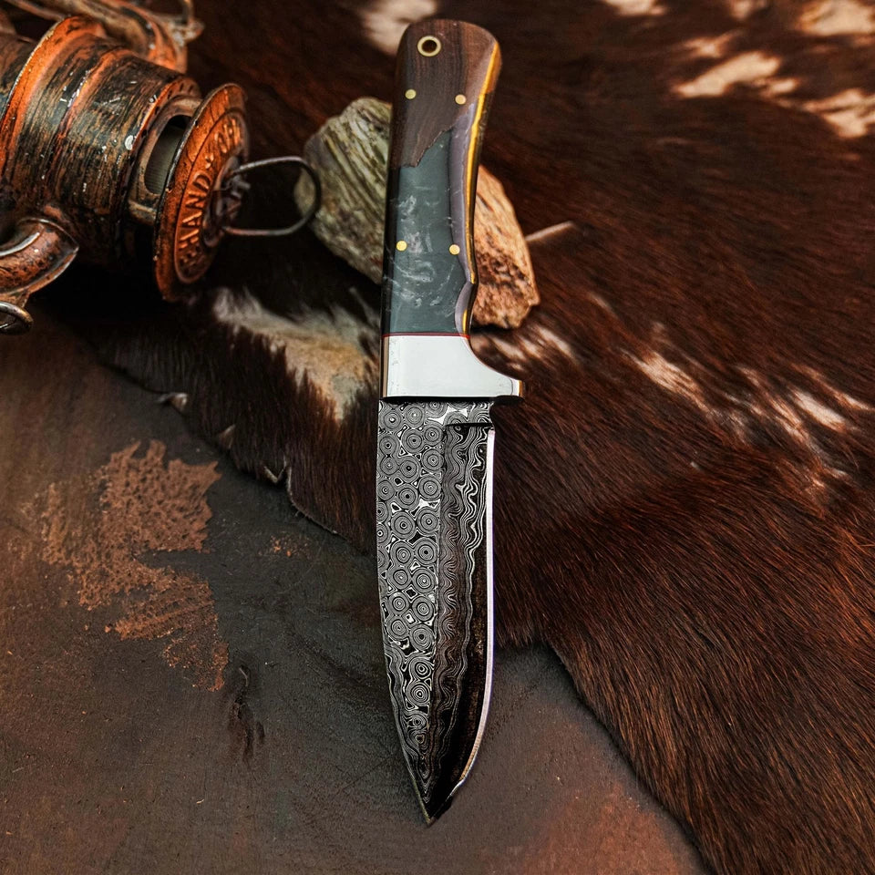 A fixed-blade knife with a patterned Damascus steel blade and a dark wood handle with brass pins, displayed on a dark textured surface with a rustic wooden spool and brown fur in the background.