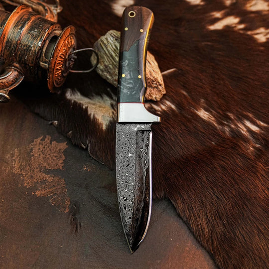 A fixed-blade knife with a patterned Damascus steel blade and a dark wood handle with brass pins, displayed on a dark textured surface with a rustic wooden spool and brown fur in the background.