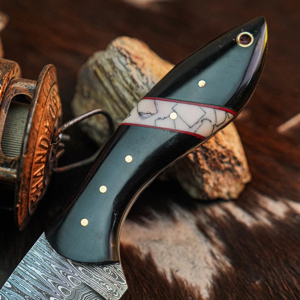 A close-up of a custom knife with a dark green handle featuring a white and gray inlay band with red trim, gold dots, and a Damascus steel blade, resting on a rustic background with a spool of thread.