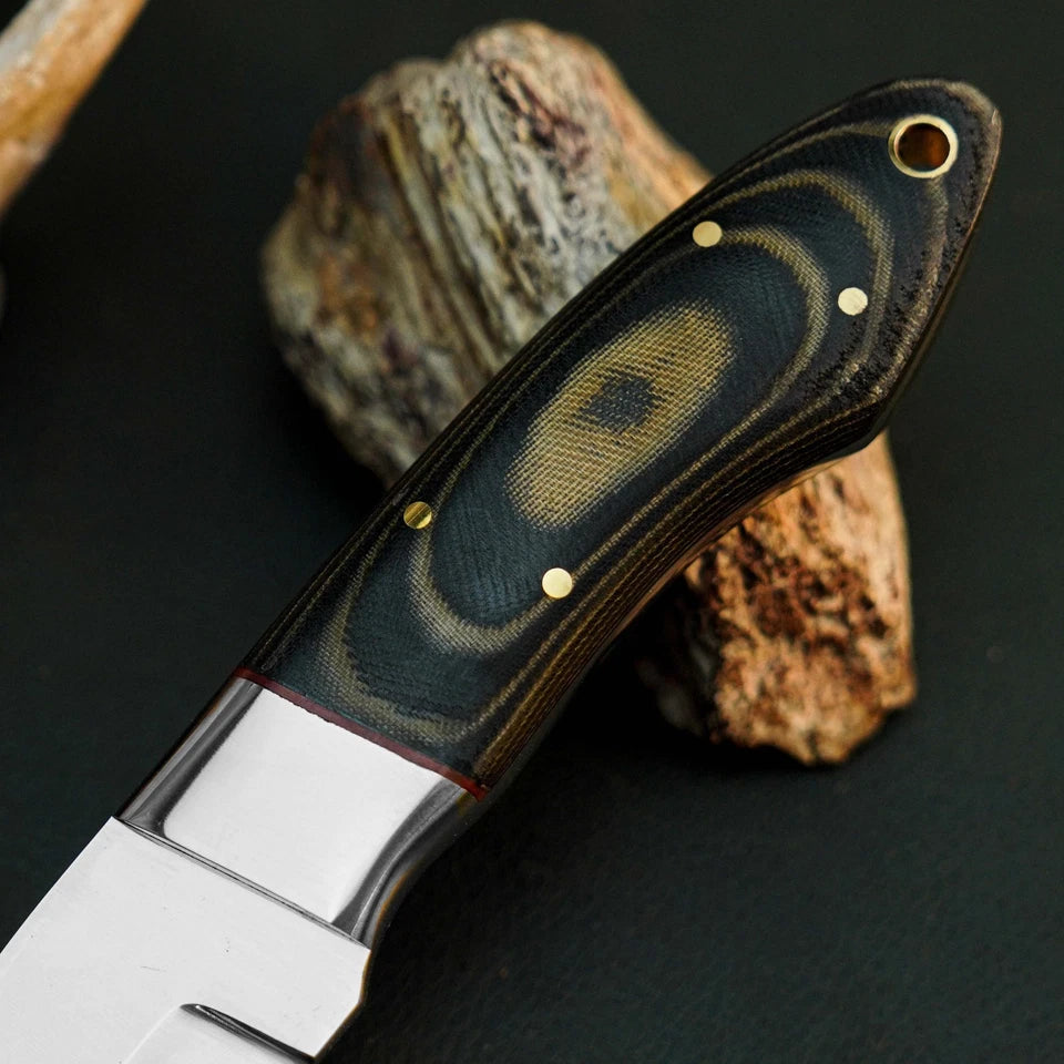 A close-up of a knife handle made from dark wood with swirling tan and black grain, accented by brass pins and a brass eyelet, resting on a dark surface with a piece of wood behind it.